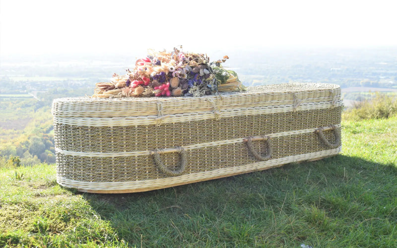 Ecoffins Pandanus Round wicker coffin with flowers on a grassy hillside