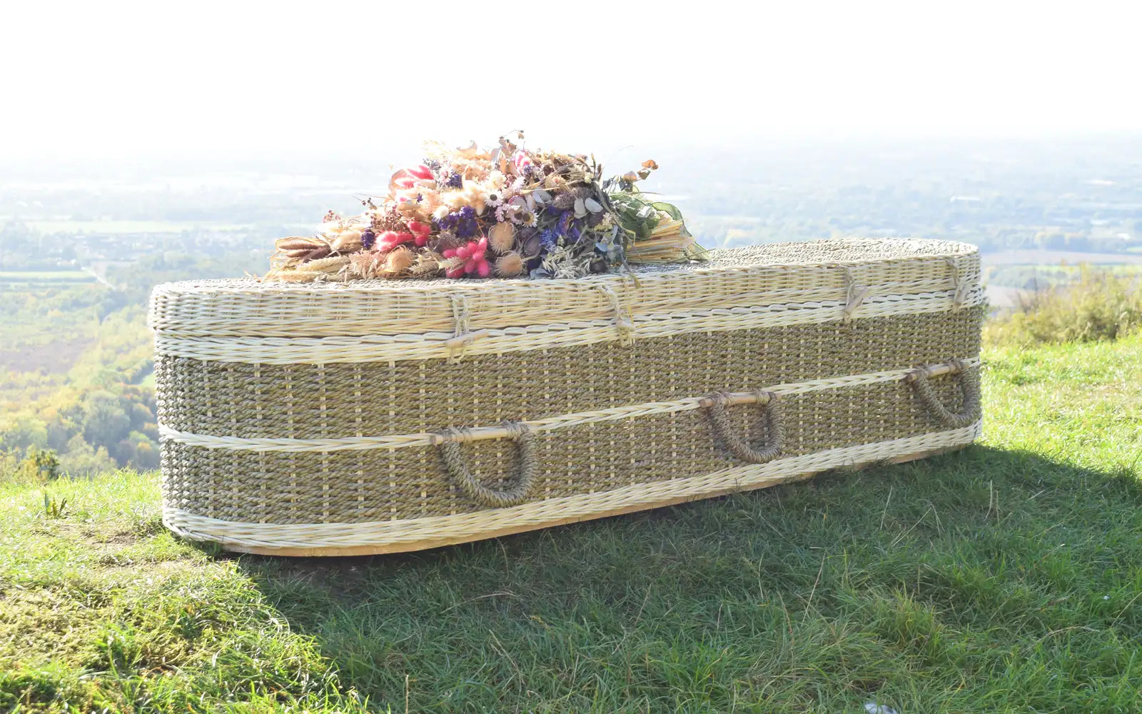 Ecoffins Pandanus Round wicker coffin with flowers on a grassy hillside