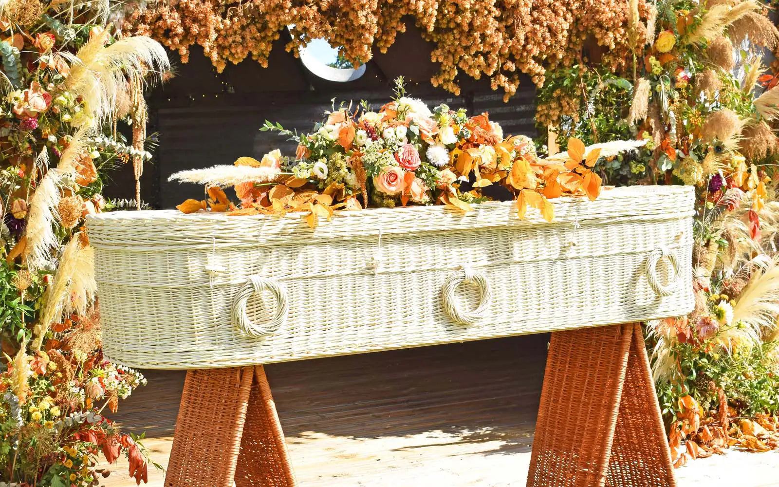 Ecoffins Willow Cromer Light Round wicker woven coffin with flowers in front of a dried flower display